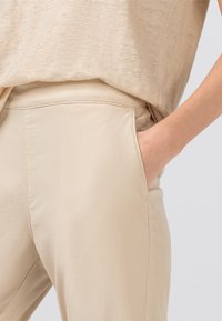 Beige lehké kalhoty s hladkým texturem, s bočními kapsami a úzkým střihem, zobrazené spolu s odpovídajícím béžovým topem.