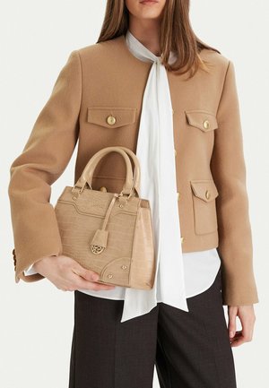 Vrouw met een jas in kamelkleur met gouden knopen, een witte blouse met een gestrikte kraag, een donkere broek en een beige handtas met textuur in haar hand.