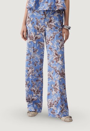 Pantaloni blu a gamba larga con grande stampa floreale marrone e bianca, indossati con pantofole beige borchiate e braccialetti d'argento su un polso.