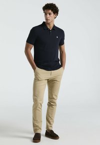 Polo shirt bleu marine à manches courtes et col, associé à un pantalon beige clair et des mocassins foncés. Détail de logo subtil sur la chemise.