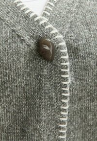 Cardigan grigio in knit con un tessuto spesso e testurizzato, un solo bottone scuro e cuciture bianche a contrasto lungo l'apertura.