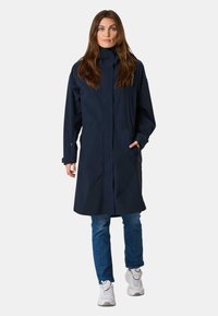 Cappotto impermeabile blu navy con colletto alto, maniche lunghe, due tasche laterali e vestibilità comoda, abbinato a jeans blu e sneakers bianche.