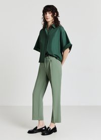 Camicia verde a maniche corte con bottoni e tasca, abbinata a pantaloni su misura verde chiaro e mocassini neri. Taglio e design semplici e moderni.