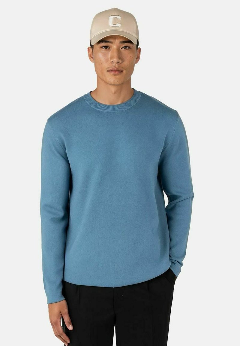 Lyseblå genser med rund hals i et mykt, teksturert strikk, kombinert med en beige caps med et "C"-logo. Modellen står mot en ensfarget bakgrunn.