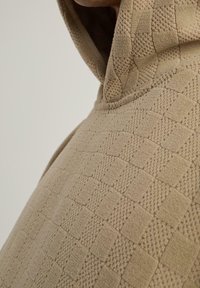 Gros plan d'une personne portant un sweat à capuche beige texturé avec un motif géométrique en relief et une couture visible le long du bord de la capuche.