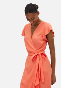 Vestido envolvente coral con mangas cortas tipo volante, escote en V profundo y un cinturón anudado en la cintura. Tejido suave con un detalle de falda drapeada.