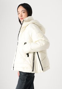 Pūdera jakas gaiši krēmkrāsā, ar kapuci, melniem rāvējslēdzējiem un teksturētu, quiltēta dizaina. Logo uzlīme uz kreisā piedurknes.