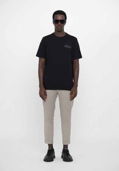 Czarny bawełniany t-shirt z kolorowym logo na piersi, w zestawieniu z jasnobeżowymi spodniami o tapered kroju i czarnymi sneakerami. Dodatki to ciemne okulary przeciwsłoneczne.