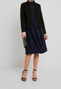 Svart skräddarsydd kavaj över en mörkblå spetsblus och en knälång kjol. Beiga ankelsandaler med rem och en blank svart kuvertväska. Slät tygtextur.