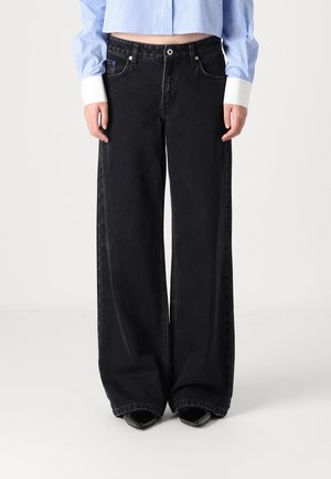 Person iført højtaljede, vidde sorte jeans og en kort blå- og hvidstribet skjorte med hvide manchetter, stående på en hvid gulv.