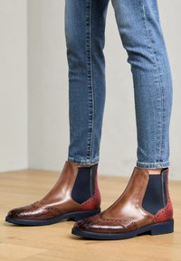 Bottines Chelsea en cuir marron avec un panneau latéral élastique bleu, présentant des détails de brogue et un capuchon de pied texturé en marron foncé. Semelle plate noire.