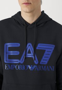 Felpa nera con cappuccio caratterizzata da una grande grafica blu con il testo "EA7 EMPORIO ARMANI". Ha un cappuccio con coulisse e una texture morbida.