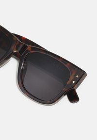 Lunettes de soleil en écaille de tortue avec verres foncés, monture carrée, légère texture et accents dorés sur les branches. Design élégant et moderne.