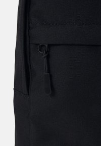 Gros plan sur une poche de sac à dos en tissu noir avec une tirette de fermeture éclair noire munie d'une boucle en corde sur un matériau texturé.