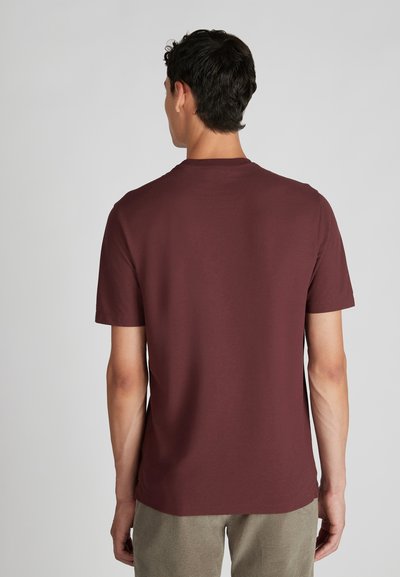 Homme avec des cheveux courts et foncés portant un t-shirt simple à manches courtes bordeaux et un pantalon beige, vu de dos sur un fond neutre.