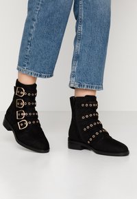 Botines de ante negros con detalles en ojetes dorados y correas con hebilla, suela plana y cremallera lateral para un fácil uso, combinados con unos vaqueros azules.