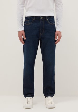 Hombre vistiendo jeans rectos azul oscuro, camisa blanca de manga larga y zapatillas blancas, posando contra un fondo liso.