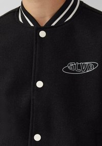 Giacca varsity nera in lana con bottoni a pressione bianchi, colletto a righe bianche e nere e logo ricamato "SLVR" sul lato sinistro del petto.