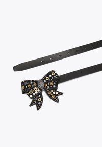 Zwarte leren riem met een strikvormige bevestiging versierd met gouden en zilveren studs, met een gestructureerd oppervlak en een metalen sluiting.