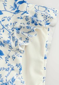Tissu blanc avec motifs floraux et feuillages bleus, présentant des bords froncés en couches et une texture douce et fluide.
