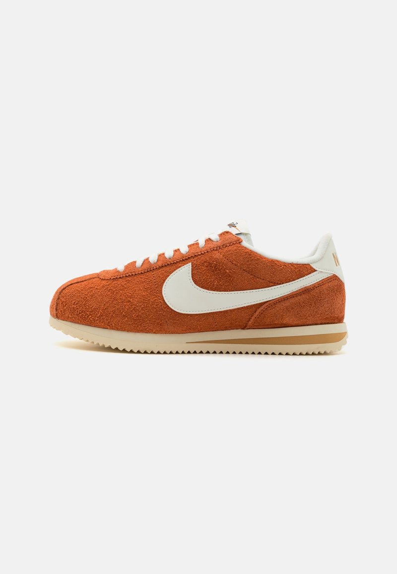 Baskets basses en daim orange avec le swoosh blanc de Nike, lacets blancs et semelle beige sur fond blanc uni.