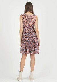 Femme portant une robe fleurie sans manches, longueur genou, avec ourlet à volants et bottines claires, debout, dos tourné, sur fond uni.