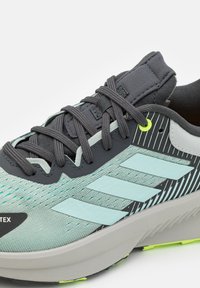 Zapatilla deportiva ligera con una parte superior de color verde menta texturizada, acentos en gris oscuro, rayas reflectantes y una suela gris acolchada.