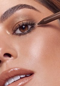 Matita eyeliner marrone applicata all'occhio di un modello, con ombretto dorato caldo, pelle chiara e colore delle labbra neutro. Vista ravvicinata.