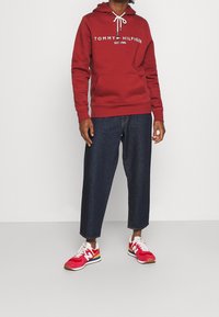 Persona vistiendo una sudadera roja de Tommy Hilfiger, jeans cortos oscuros y zapatillas rojas de New Balance, de pie contra un fondo sencillo.