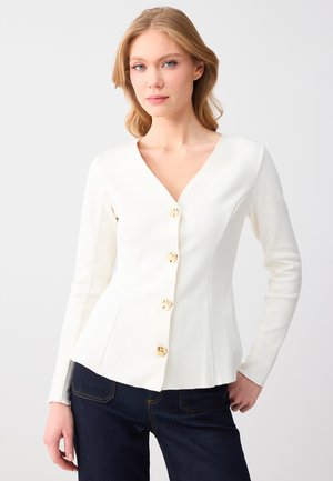 Donna con capelli biondi che indossa un top bianco aderente a maniche lunghe con bottoni dorati e jeans blu scuro, in piedi contro uno sfondo chiaro e uniforme.