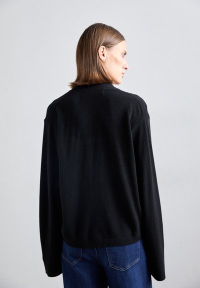 Sweater preto oversized tricotado com decote redondo e mangas longas. Textura suave e bainha inferior canelada. Usado com jeans de ganga azuis.