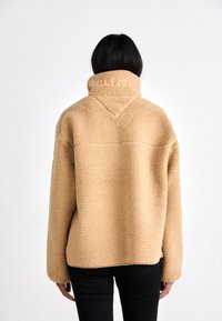 Person with black hair wearing a beige sherpa fleece jacket with high collar and black pants, seen from the back on a white background.