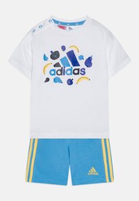 Camicia bianca a maniche corte con grafiche di frutta colorate e logo "adidas"; abbinata a pantaloni corti blu con strisce gialle e dettaglio del logo.