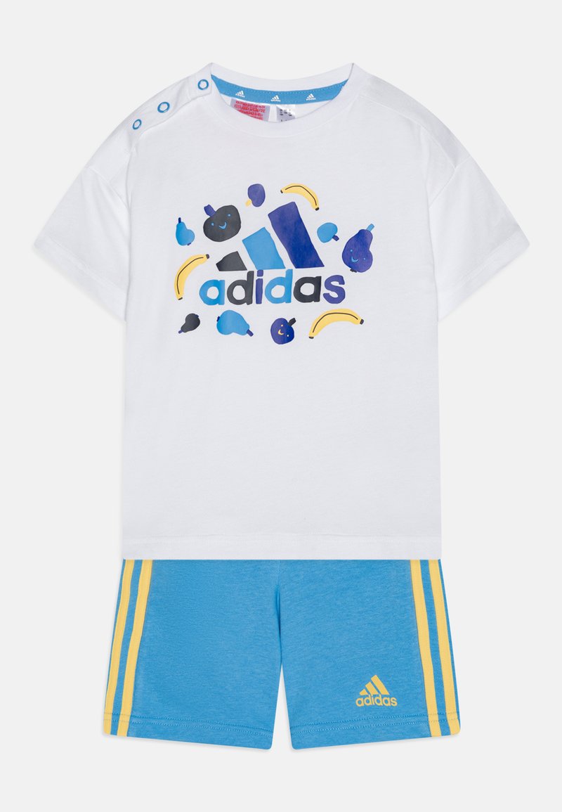 Camicia bianca a maniche corte con grafiche di frutta colorate e logo "adidas"; abbinata a pantaloni corti blu con strisce gialle e dettaglio del logo.