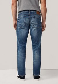 Hombre vistiendo jeans rectos desgastados de color azul y zapatillas negras, de pie de espaldas contra un fondo claro y liso.