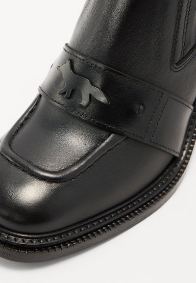 Bota de cuero negro con acabado suave, punta redondeada y una correa decorativa con un emblema de zorro. Detalles de costura a lo largo de la suela.