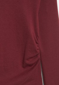 Burgunderrote Tunika mit weicher Textur, mit gerafftem Seitendetail und Seitenschlitz, aus einem dehnbaren Baumwollmischgewebe gefertigt.