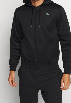 Mann trägt einen schwarzen Zip-up-Hoodie mit einem kleinen grünen Krokodil-Logo auf der Brust und passende schwarze Hose vor einfarbigem Hintergrund.
