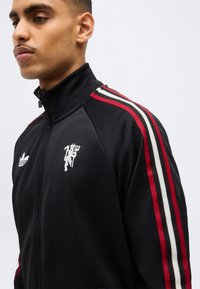 Νεαρός άνδρας που φοράει μαύρο αθλητικό μπουφάν με φερμουάρ, με ρίγες κόκκινες, λευκές και μαύρες στα μανίκια και λευκά λογότυπα στο στήθος.