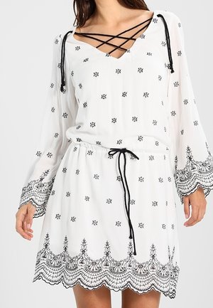 Valkoinen mekko, jossa mustat kirjailut, pitkät hihat, V-kaula-aukko ja nyöritys. Kiristysnauha vyötäröllä; aaltoileva helma.