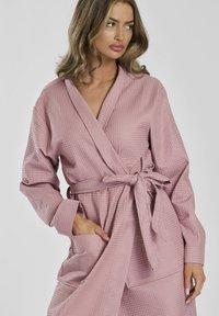 Accappatoio rosa chiaro, strutturato, con design avvolgente, maniche lunghe e cintura annodata. Presenta una tasca frontale e una finitura in tessuto morbido.