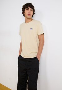 T-shirt beige à manches courtes avec un petit logo bleu marine sur la poitrine. Associé à un pantalon noir, porté avec les mains dans les poches.