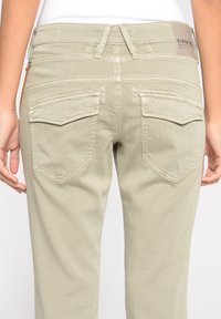 Ljus khaki åtsittande byxor med två bakfickor, synlig sömnad och en märkesläderdetalj vid midjan. Slät textur.