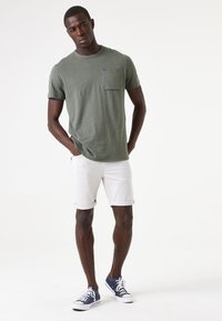 Olivgrünes Baumwoll-T-Shirt mit einer Brusttasche, kombiniert mit hochgerollten weißen Jeansshorts und navyblauen Segeltuchschuhen.