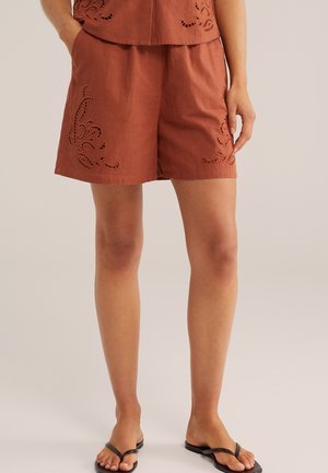 Roostevärvi lühikesed püksid lillemustriliste kirjatud lõigetega, kantud sobiva ülaosaga ja mustade varbavahekingadega, modell seisab vasak käsi taskus.