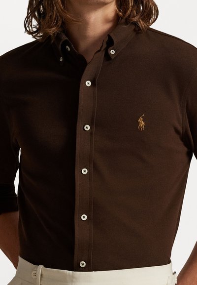 Καστανό πόλο μπλουζάκι με υφαντή υφή, διαθέτοντας κολάρο με κουμπιά, λευκά κουμπιά και κεντημένο λογότυπο στο στήθος.