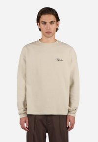 Beige waffle-strikket langærmet bluse med rund hals og broderet logo på venstre bryst. Løs pasform med ribbede manchetter.