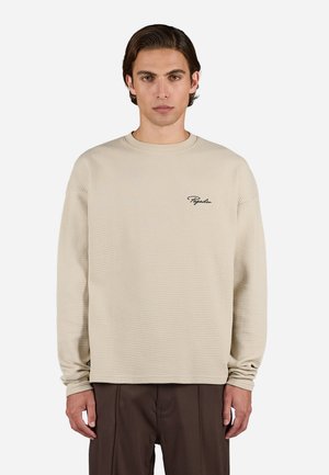 Beige waffle-strikket langærmet bluse med rund hals og broderet logo på venstre bryst. Løs pasform med ribbede manchetter.