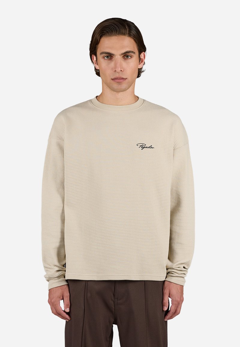 Beige waffle-strikket langærmet bluse med rund hals og broderet logo på venstre bryst. Løs pasform med ribbede manchetter.