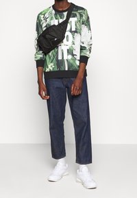 Felpa a maniche lunghe con motivo floreale verde e testo bianco; abbinata a pantaloni in denim scuri e sneakers bianche, con una borsa a tracolla nera.